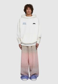 Vit hoodie med framficka, med blå och svart text. Matcher med vida byxor som graderar från rosa till lila. Vita sneakers.