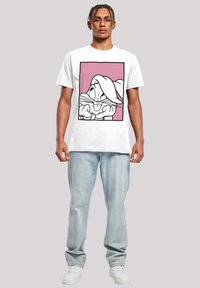 Hvit bomulls t-skjorte med en svart omriss tegneseriefigur på rosa bakgrunn. Modell har på seg lyseblå jeans og hvite joggesko.
