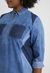 Camicia di jeans azzurro chiaro con zip frontale, due tasche sul petto e inserti in denim blu scuro. Maniche arrotolate con una trama morbida.