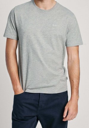 Uomo con t-shirt grigio chiaro a maniche corte e pantaloni blu scuro, con una mano in tasca e l'altra rilassata lungo il fianco.
