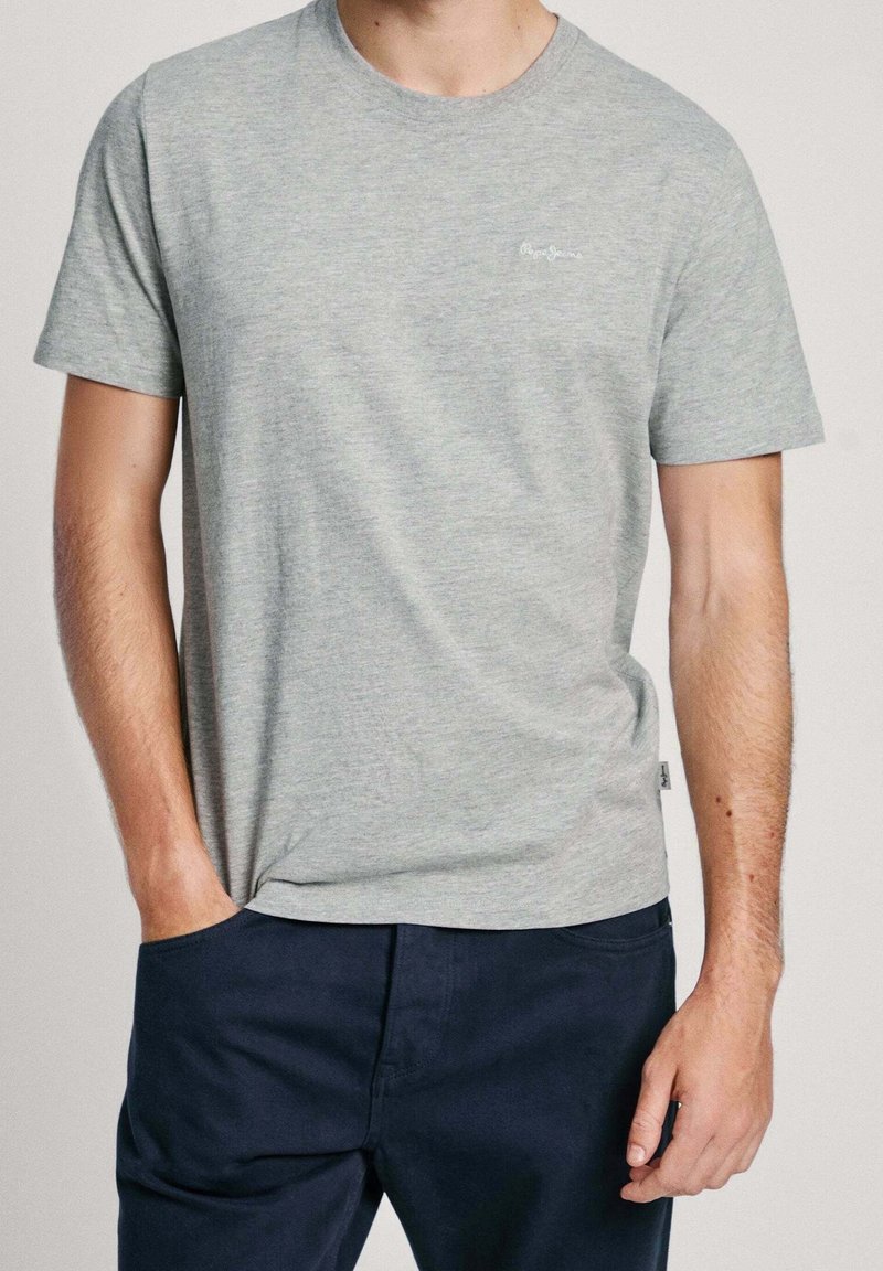 Homme portant un t-shirt gris clair à manches courtes et un pantalon bleu foncé, une main dans la poche et l'autre détendue le long du corps.