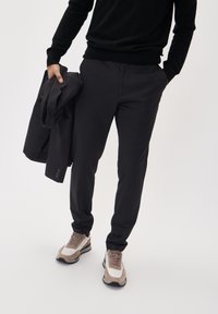 Čierne pletené nohavice so slim fit strihom, skombinované s čiernym sveterom a šedo-hnedými teniskami. V jednej ruke drží tmavý blejzer.