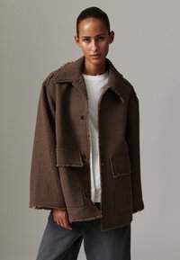 Brown, textured jacket with a collar, front pockets, and frayed edges, layered over a white shirt and paired with dark gray pants.