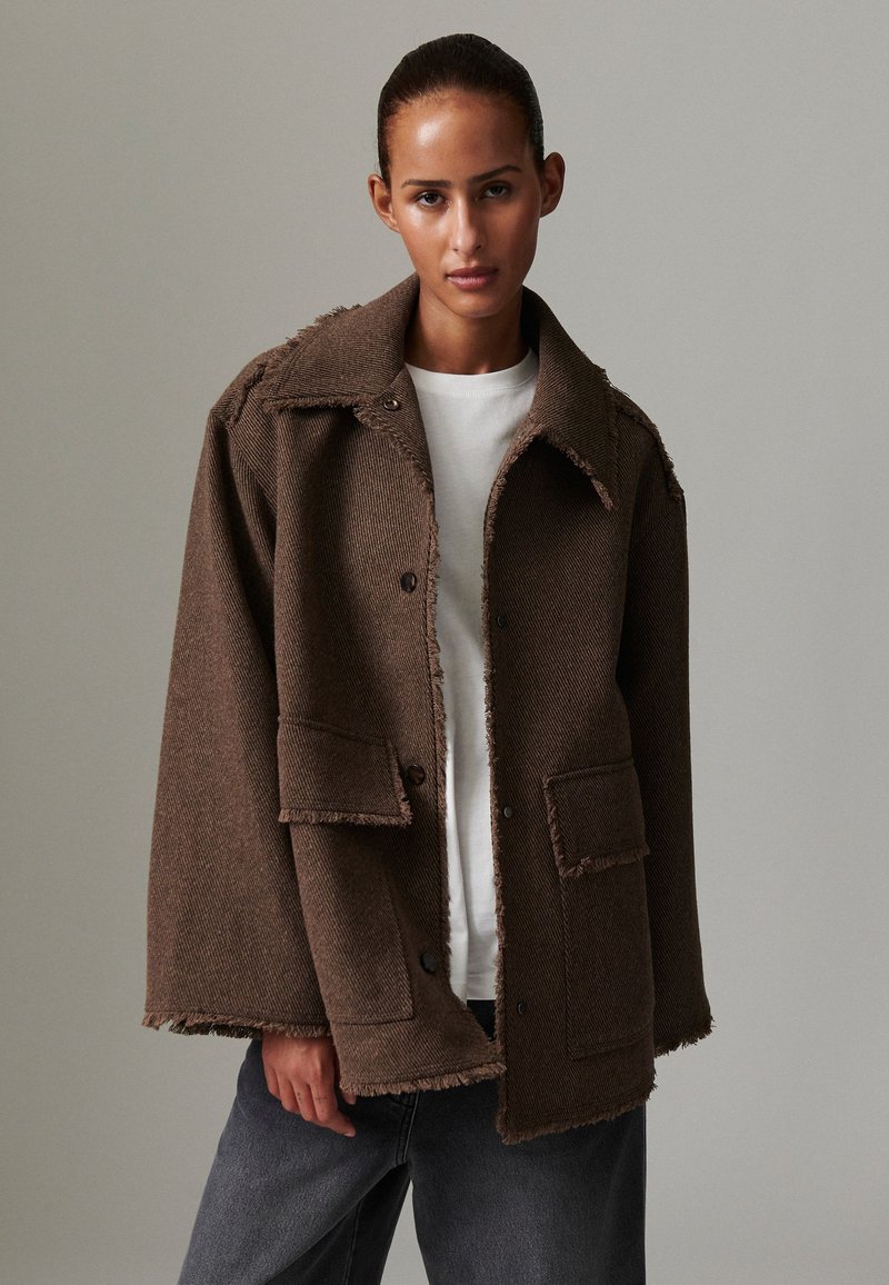 Brown, textured jacket with a collar, front pockets, and frayed edges, layered over a white shirt and paired with dark gray pants.