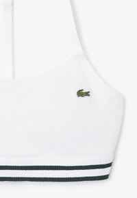 Sujetador deportivo blanco con tirantes ajustables, logo de cocodrilo verde en el lado izquierdo y banda elástica a rayas blancas y negras en la parte inferior.