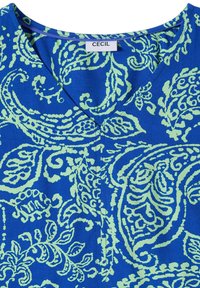 Tissu bleu avec des motifs paisley vert clair et un col en V avec une étiquette de marque "CECIL" visible.