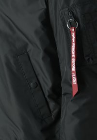 Černá nylonová bomber bunda s kapsou na zip, kulatým logem na knoflíku a červenou visačkou "ODSTRANIT PŘED LETEM" připevněnou na kroužku.