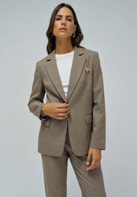 Blazer marrón a cuadros con cuello con muesca, dos bolsillos delanteros y un broche decorativo. Lleva puesto sobre una camiseta blanca. Corte ajustado.