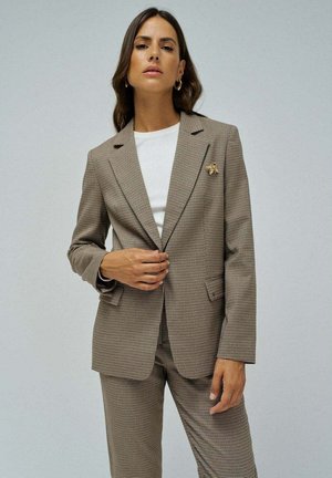 Blazer marrone a quadri con colletto a scollatura, due tasche frontali e una spilla decorativa. Indossato sopra una t-shirt bianca. Vestibilità su misura.