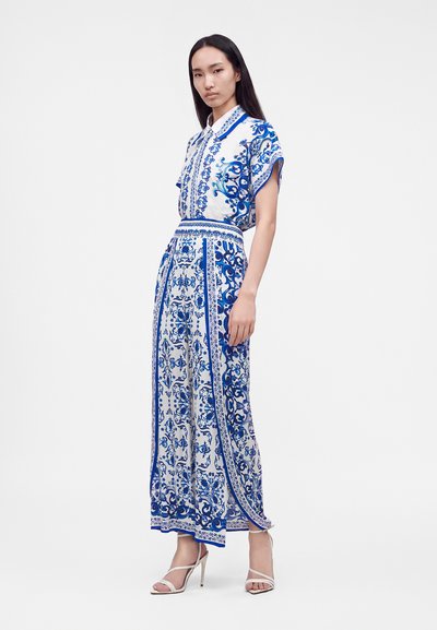 Femme portant un chemisier à manches courtes blanc et bleu à motifs et une jupe longue assortie, associée à des sandales blanches à talons hauts, debout.