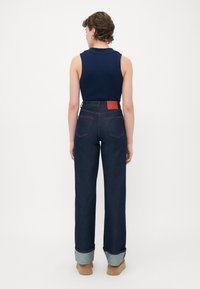 Kvinne som står med ryggen til, iført ermeløs marineblå topp, høyt midje mørkeblå jeans med oppbrettede hem, og beige platå sko mot en hvit bakgrunn.