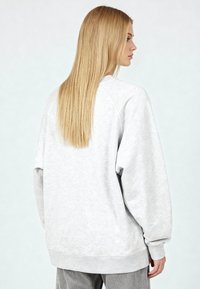 Sweat-shirt gris à coupe décontractée, manches raglan, poignets et ourlet côtelés, tissu doux et texturé. Vue de dos montrant des cheveux longs et raides.