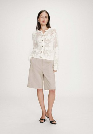 Femme debout, mains dans les poches, portant un cardigan blanc à maille ouverte, un short beige à hauteur des genoux et des sandales noires à talons.