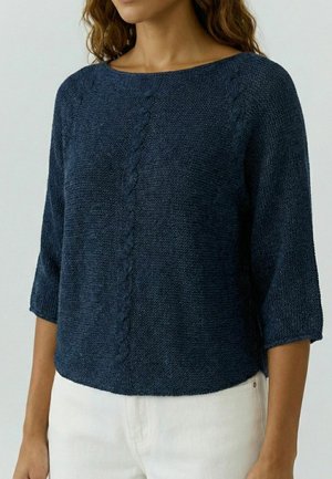 Vrouw draagt een marineblauwe gebreide trui met 3/4 mouwen en witte broek, zichtbaar van nek tot heupen.