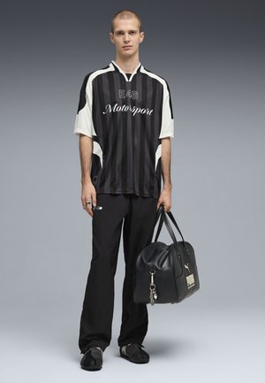 Camiseta de rayas en blanco y negro con el texto "E46 Motorsport", combinada con pantalones negros y un bolso de viaje de cuero negro con herrajes plateados.