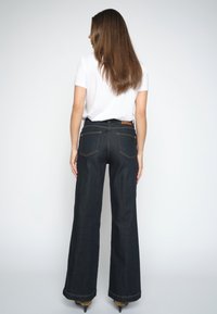Donna con capelli lunghi e castani che indossa una camicia bianca e jeans scuri a gamba larga, in piedi di spalle contro uno sfondo chiaro e uniforme.