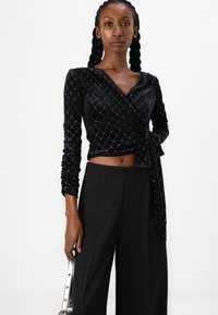 Μαύρη βελούδινη τοπ με μακριά μανίκια, με σχέδιο διαμαντιού και πλαϊνό δέσιμο. Συνδυασμένη με μαύρες παντελόνες με σιλουέτα ψηλής μέσης.