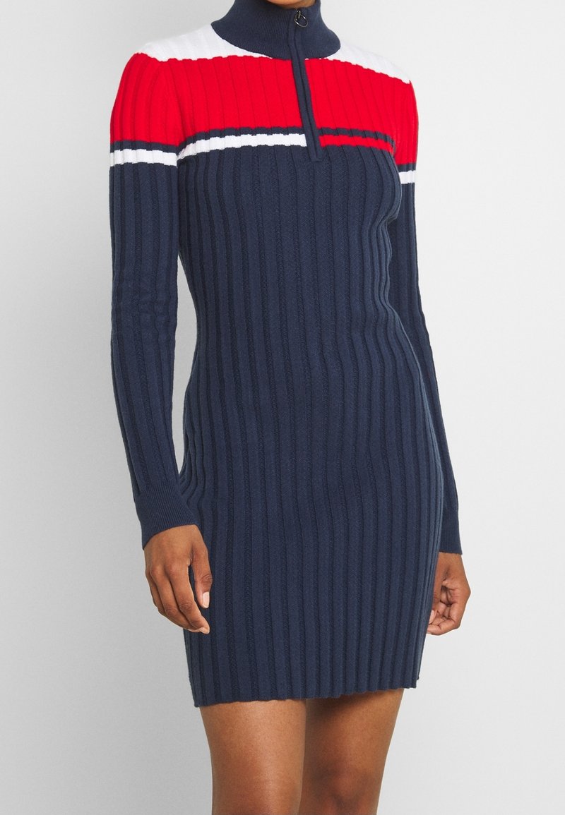 Femme portant une robe en maille côtelée bleu marine à manches longues, avec une encolure color-block rouge, blanche et bleu marine, et un col montant zippé.