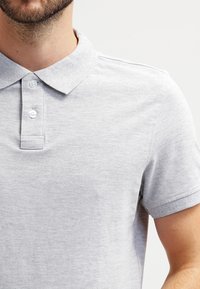Harmaa polo-paita, joka on valmistettu teksturoidusta kankaasta ja jossa on klassinen kaulus, kaksinappinen etusuljin sekä lyhyet hihat.