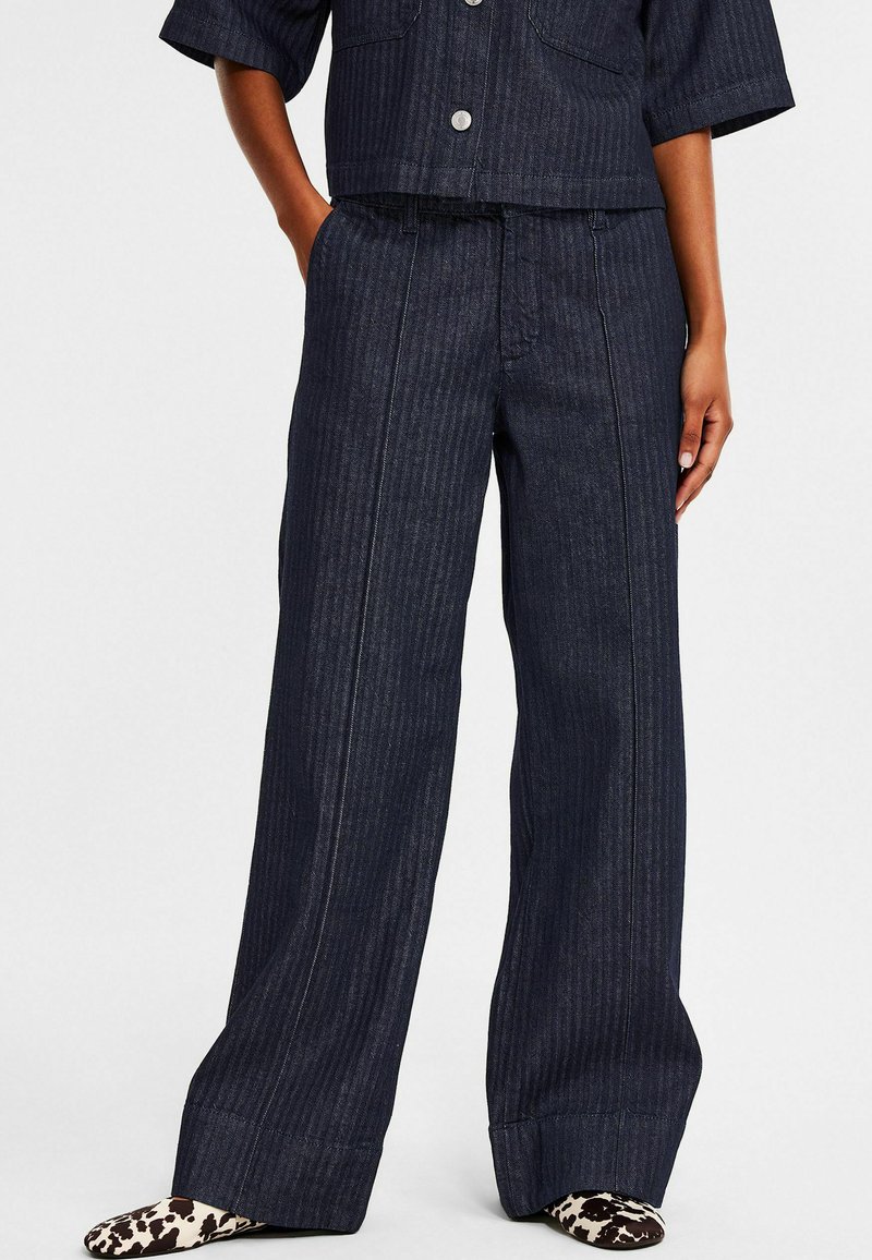 Mörk jeans med vida ben och vertikalt randmönster. Har framfickor, knappstängning och subtila sömnadsdetaljer.