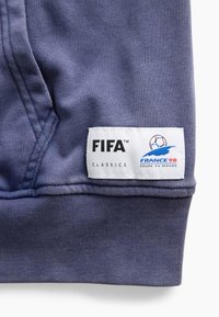 Μωβ ύφασμα σε κοντινό πλάνο με μια λευκή ορθογώνια ετικέτα που δείχνει τα λογότυπα FIFA Classics και France 98 Coupe du Monde.