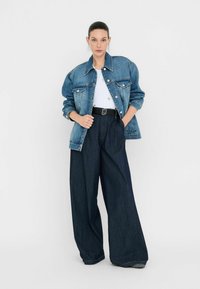 Giubbotto di jeans di colore azzurro chiaro con chiusura a bottoni e tasche a toppa, abbinato a jeans a gamba ampia di colore blu scuro e una t-shirt bianca, con una cintura.