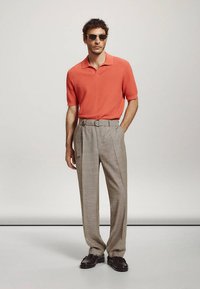 Homme portant un polo en maille corail à manches courtes, un pantalon beige taille haute avec ceinture, des chaussures noires et des lunettes de soleil foncées, debout devant un fond uni.