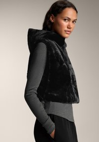 Vest negru din blană artificială, fără mâneci, cu glugă, având un guler înalt și o textură moale. Purtați peste un top lung cu mânecă lungă, de culoare gri, cu dungi.