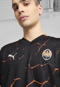 Camiseta deportiva negra con patrón de grietas naranjas, con cuello en V, un logo blanco de Puma y un escudo circular del Shakhtar Donetsk.