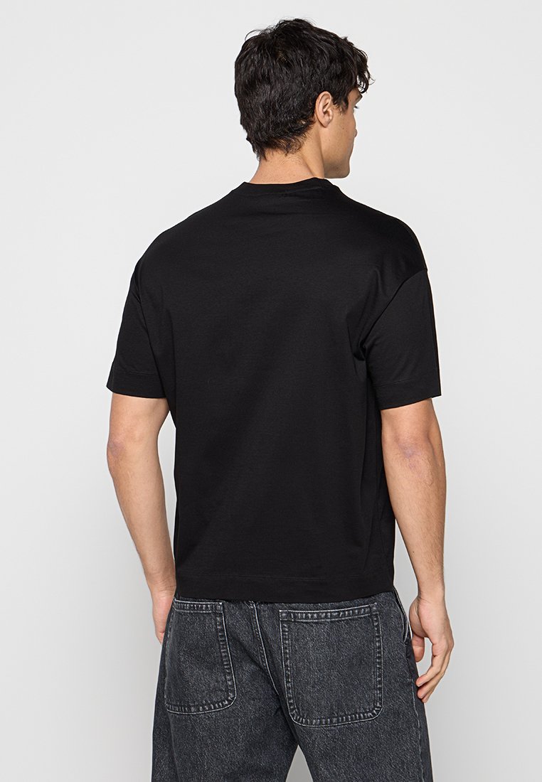 Hombre con cabello corto y oscuro, que lleva una camiseta negra de manga corta lisa y vaqueros negros descoloridos, de pie de espaldas sobre un fondo blanco liso.