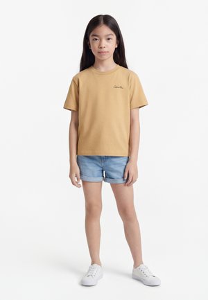 Meisje staat, draagt een beige Calvin Klein t-shirt, lichtblauwe opgerolde denim short en witte sneakers, op een witte achtergrond.