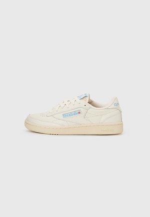 Mimo biele teniska značky Reebok s svetlomodrým logom a béžovou podrážkou, bočný pohľad na jednoduchom svetlosivom pozadí.