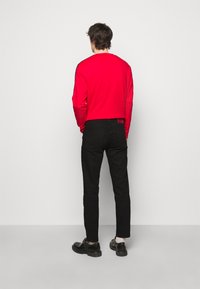 Muž stojící zády, oblečený v červené košilové dlouhé tričko, černé slim-fit kalhoty a černé formální boty s bílými ponožkami, na jednobarevném pozadí.