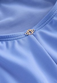 Hellblauer Stoff mit glatter Textur, auf einer Seite mit Mesh-Detail und einem goldfarbenen, runden Metallakzent in der Nähe des Halsausschnitts.