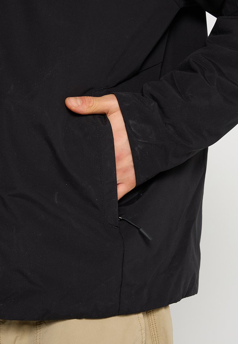 Giacca nera con una tasca con zip sul lato sinistro, realizzata in un materiale liscio e opaco. Le maniche sono leggermente larghe, la texture appare resistente.