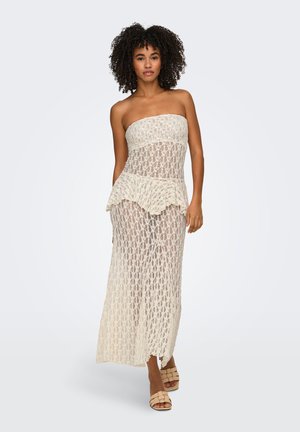 Femme aux cheveux bouclés portant une robe longue en dentelle transparente sans bretelles avec détail peplum et sandales tressées couleur nude, debout sur un fond uni.
