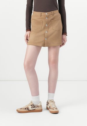 Khaki mini nederdel med frontlukning med knapper, to forlommer og en tætsiddende design. Kombineret med mønstrede sneakers og hvide ankelstrømper.