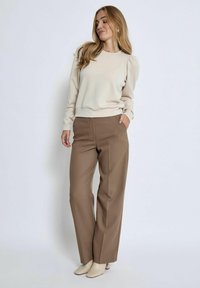 Beżowy sweter z bufkami, zestawiony z brązowymi spodniami o wysokim stanie. Model ma na sobie kremowe szpilki. Prosty, klasyczny design o czystych liniach.
