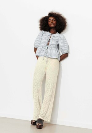 Femme aux cheveux bouclés volumineux portant un chemisier rayé avec des liens et un pantalon large texturé couleur crème, debout contre un mur blanc.