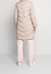 Abrigo puffer de un beige claro con costuras en forma de chevrón, diseño hasta la rodilla, puños acanalados y un cuello alto, combinado con pantalones rosa y zapatillas blancas.