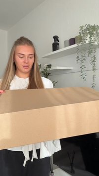 Cutie de carton ținută de o persoană, formă dreptunghiulară, culoare maro deschis, suprafață netedă. În fundal se văd rafturi cu plante și sticle.