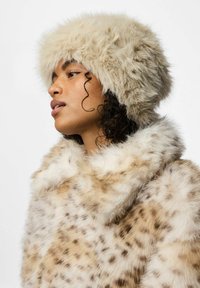 Cappello in finta pelliccia di colore crema con una parte superiore pelosa. La modella indossa un cappotto in finta pelliccia a macchie, caratterizzato da una texture morbida e un colletto arrotondato.