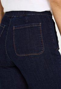 Pantalones vaqueros de mezclilla azul oscuro con diseño ajustado, cinturilla elástica y un bolsillo trasero con detalles de costuras naranjas.