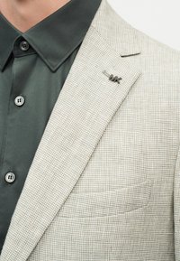 Záber zblízka na osobu nosiacu svetlo béžové kárované sako s kovovou pripínacou brošňou "MK" na klopne a tmavozelenú košeľu na gombíky.