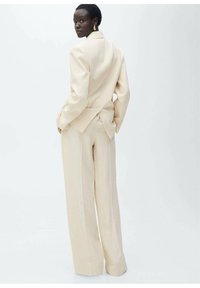 Beige räätälöity puku, jossa on selkeä rakenne, laajalahkeiset housut ja blazer, jossa on lovikaulus ja takana ilmestyvä venttiili.