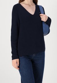 Femme portant un pull bleu marine à col en V et un jean bleu, tenant un sac à main bleu sur son épaule.