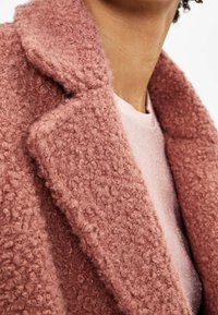 Cappotto rosa testurizzato e peloso con colletto a lancia, esposto accanto a un top in maglia con scintillio rosa chiaro. Vista ravvicinata del tessuto e dei dettagli.