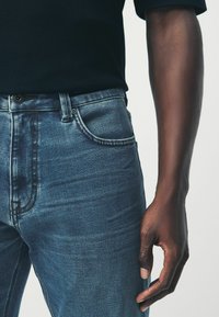 Avant-bras droit d'une personne portant une chemise noire et un jean en denim bleu sur un fond clair.
