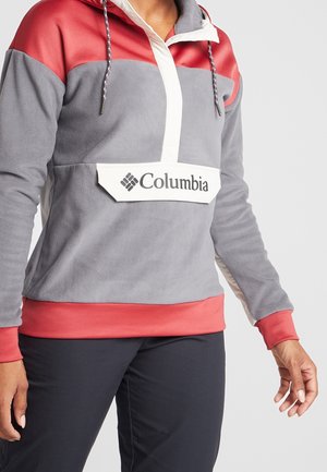 Personne portant un pull polaire Columbia gris et rouge avec des accents blancs et un pantalon noir, visible du cou aux hanches.