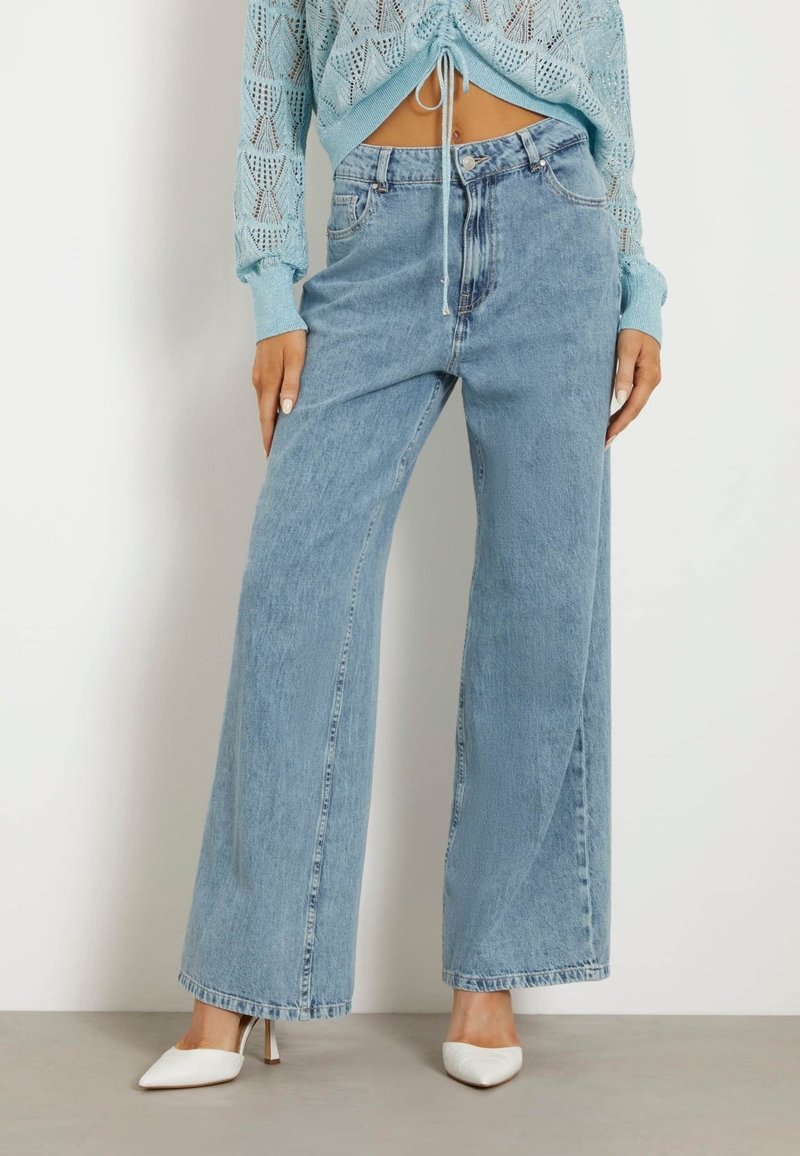 Jean bleu clair à jambes larges associé à des chaussures blanches à talons et un pull en maille bleu court à manches longues avec un cordon à l'avant.